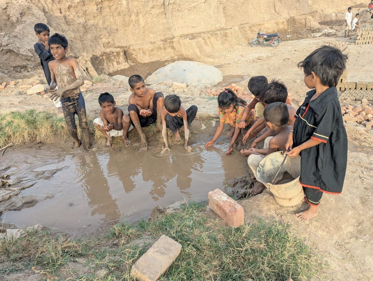 Children gathered at a muddy water source near a brick kiln